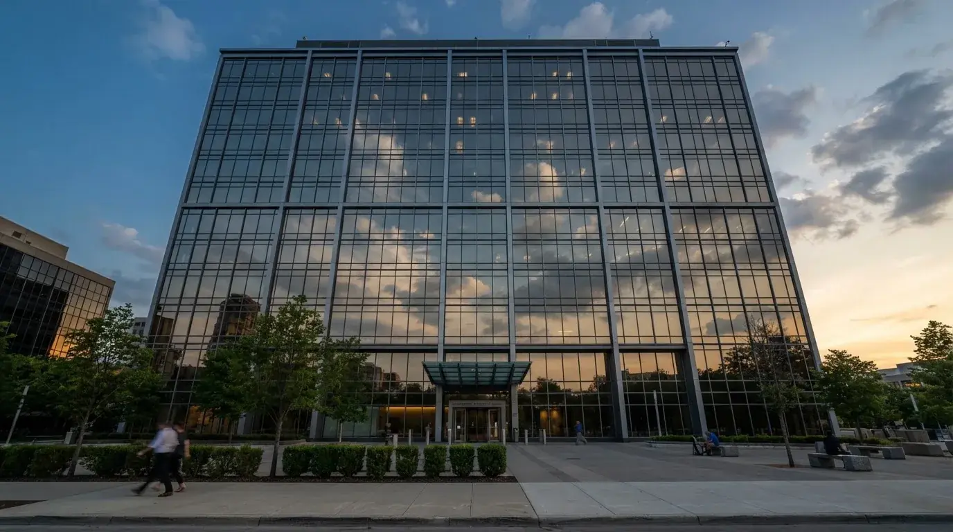 Un edificio de oficinas con fachada de cristal reflejando nubes, fotografiado desde una acera ancha por la tarde.